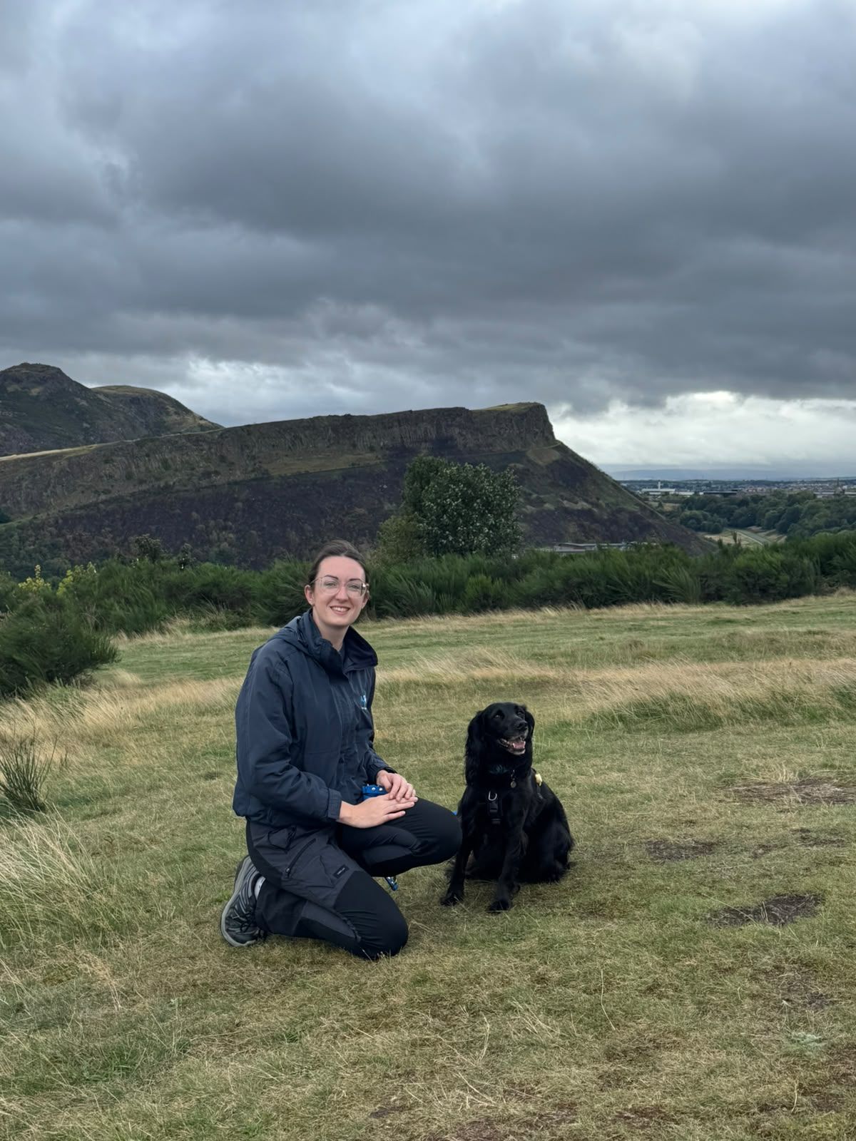 Rosy kneeling beside her black spaniel in Holyrood Park, with Arthur’s Seat behind them
