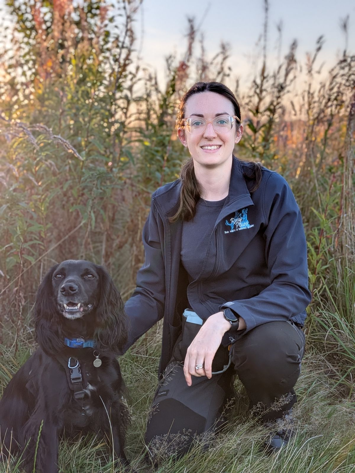 Rosy smiling and kneeling beside her black spaniel in long grass at sunset